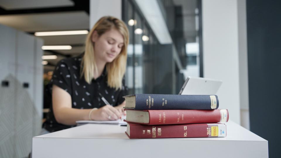 Woman sat in the library writing with a stack of books next to her