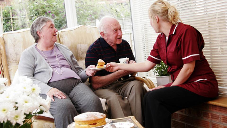 Two older people having tea and cake with their carer in a care home