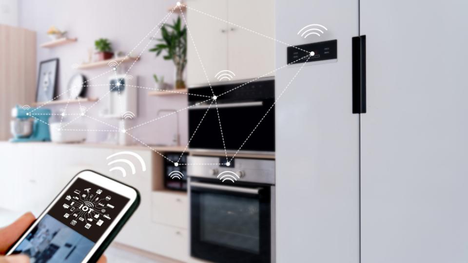 A phone that has the letters IOT displayed at the screen is shown alongside a fridge and an oven. There are dotted lines between all of the devices to show that they are connected.