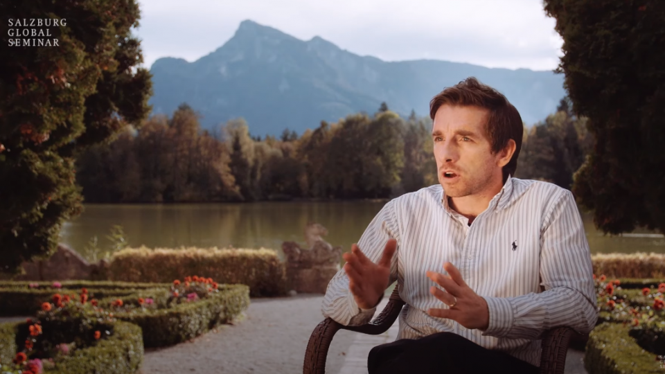 Graeme sits in a garden by a lake in front of mountains to conduct an interview with a Salzburg media centre
