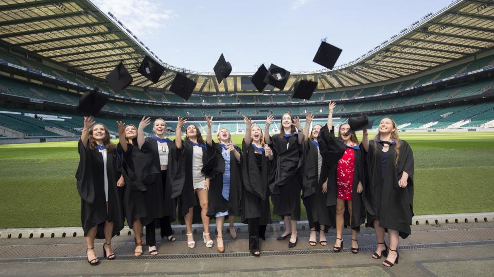 A group of graduates throwing their mortarboard hats in the air at Twickenham Stadium.