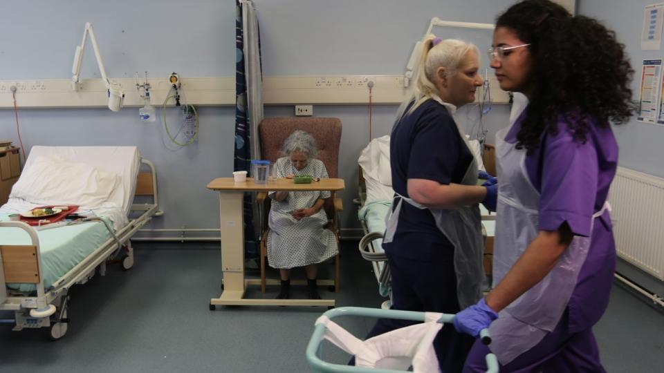 Woman in hospital chair on ward with nurses walking past