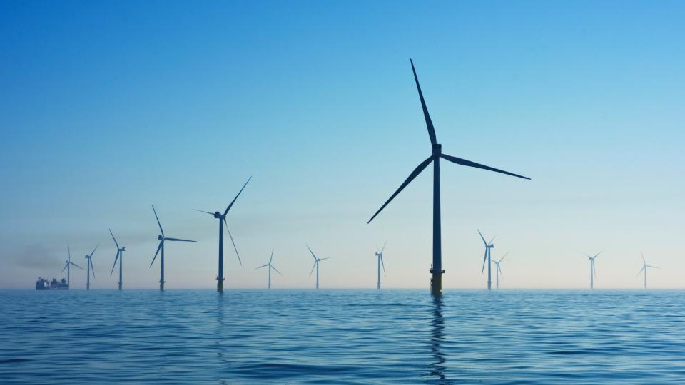 Offshore wind turbines are pictured rising out of a blue ocean. The sky is clear blue and the turbines can be seen disappearing into the horizon.