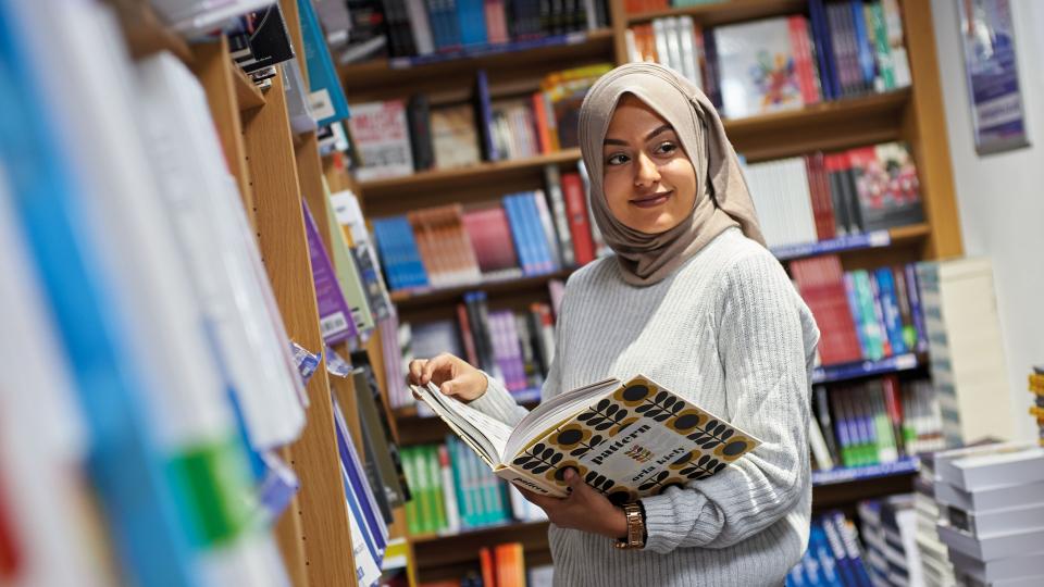 POC student wearing a grey hijab, is holding a book on 'pattern' in the library.