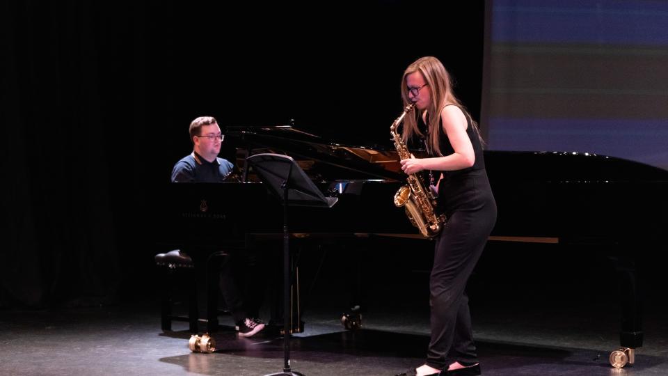Two LCM musicians performing. One is playing a grand piano and the other is playing a saxophone. Both musicians are wearing all black.