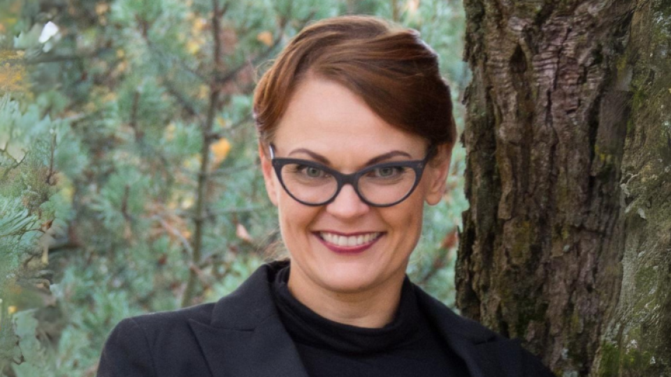 Tiina Vaittinen has short brown hair, and is wearing glasses and a black top and jacket.