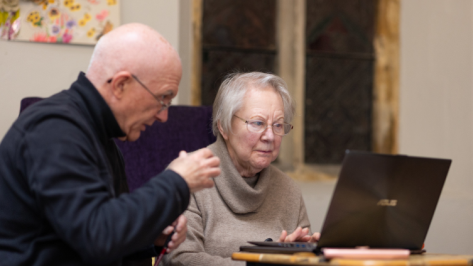 Elderly lady seated in front of a laptop assisted by someone next to her.
