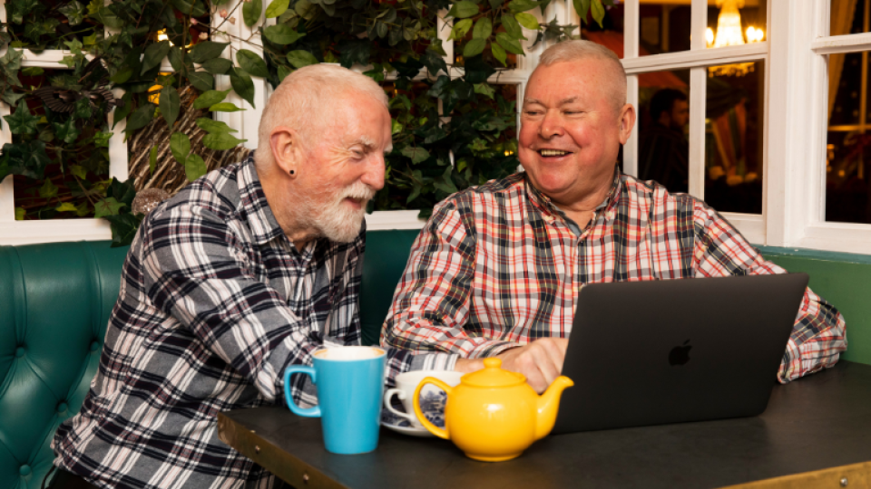 Two men sat in front of a laptop socialising and smiling.