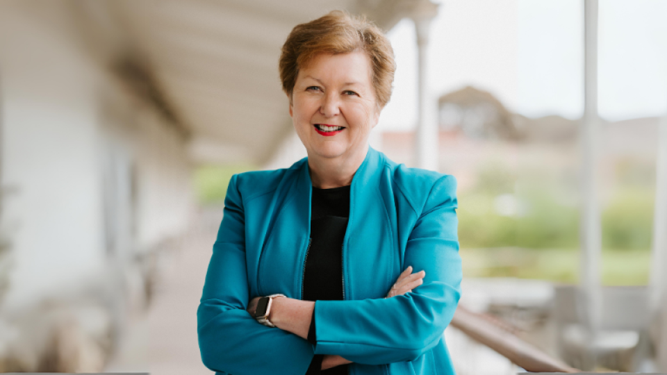 Joan Ostaszkiewicz is smiling and wearing a turquoise suit jacket and black shirt and is crossing her arms.