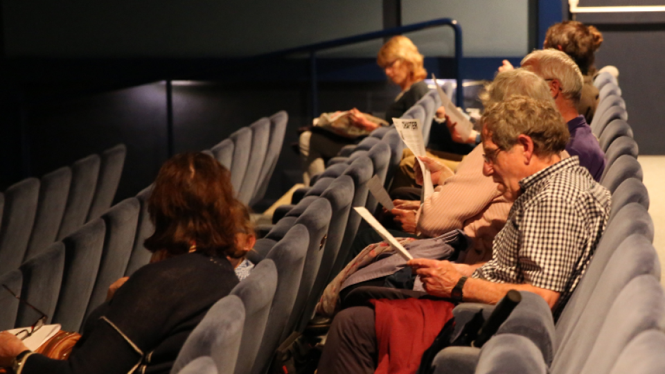 A group of adults seated at a cinema screening, reading while they wait.