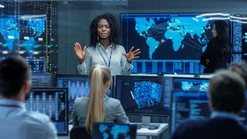 A female talking to colleagues in front of large data monitors