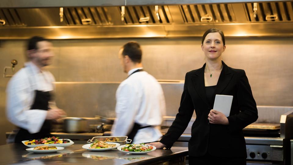 A smartly dressed woman in a busy kitchen near two chefs and plates of food
