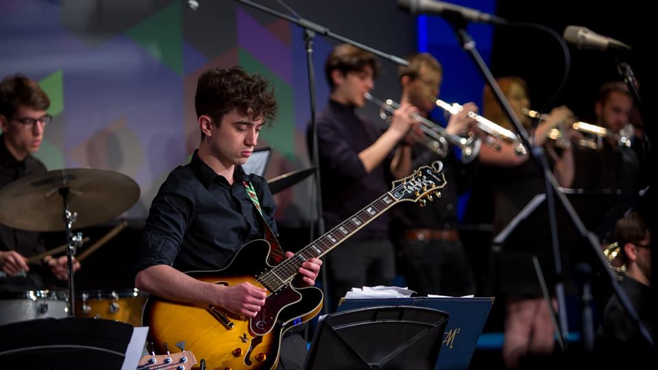 A male student on a guitar with a brass section in the background during a live performance