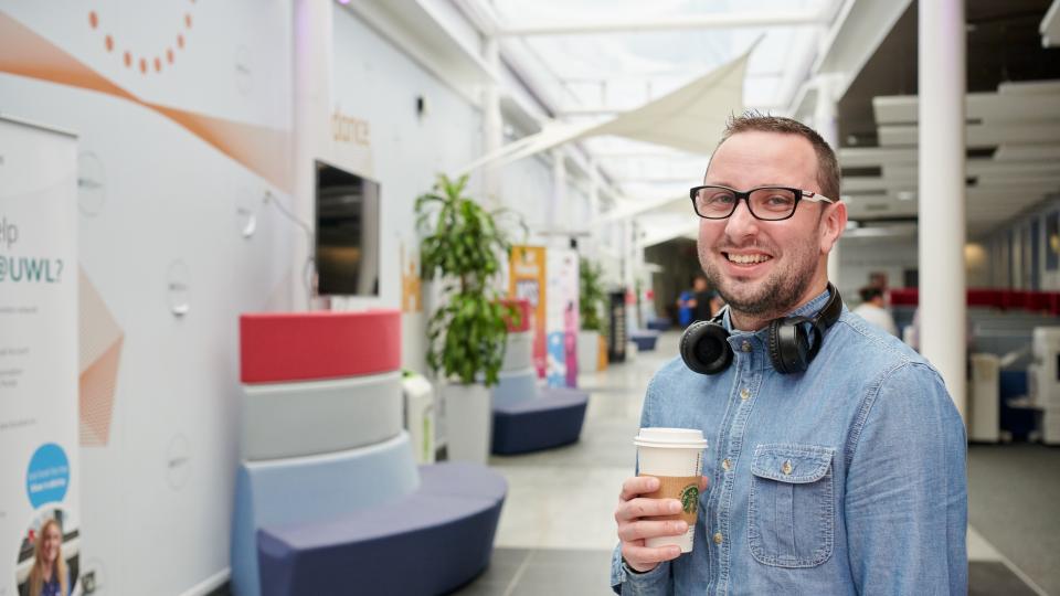 Man with glasses and head phones holding a cup of coffee smiling