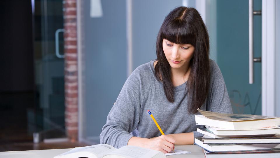 A female student with long dark brown hair is studying with several textbooks