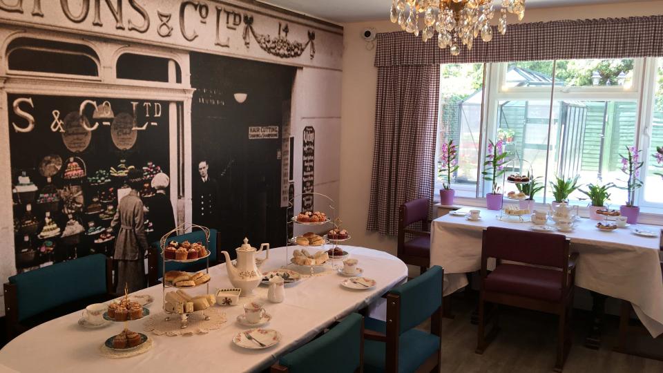 The tea room in Devonshire dar care centre, In the foreground there is a table set up for afternoon tea, in the background there is a wall designed to look like a tea shop front with the words "Lyons and Co Ltd". 