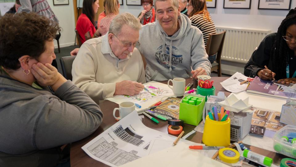 An older man is drawing using coloured pens at a table that is covered in art supplies. In the backgrounds there are other tables with different people sat at them.