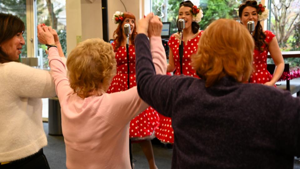 In the background, three women are singing in polka-dot dresses into retro microphones. In the foreground, woman with their backs turned to the camera dance.