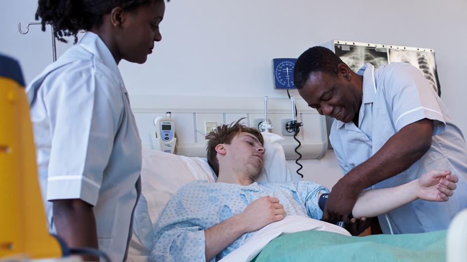 Male nurse tending to patient in hospital bed, with a second nurse present.