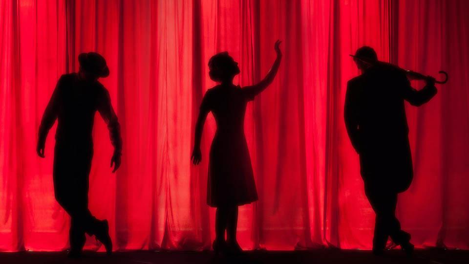 Three actors on stage silhouetted in front of a red curtain