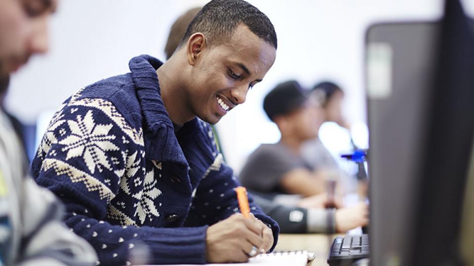 A student in a blue and white jumper writes in a notepad in front of a computer