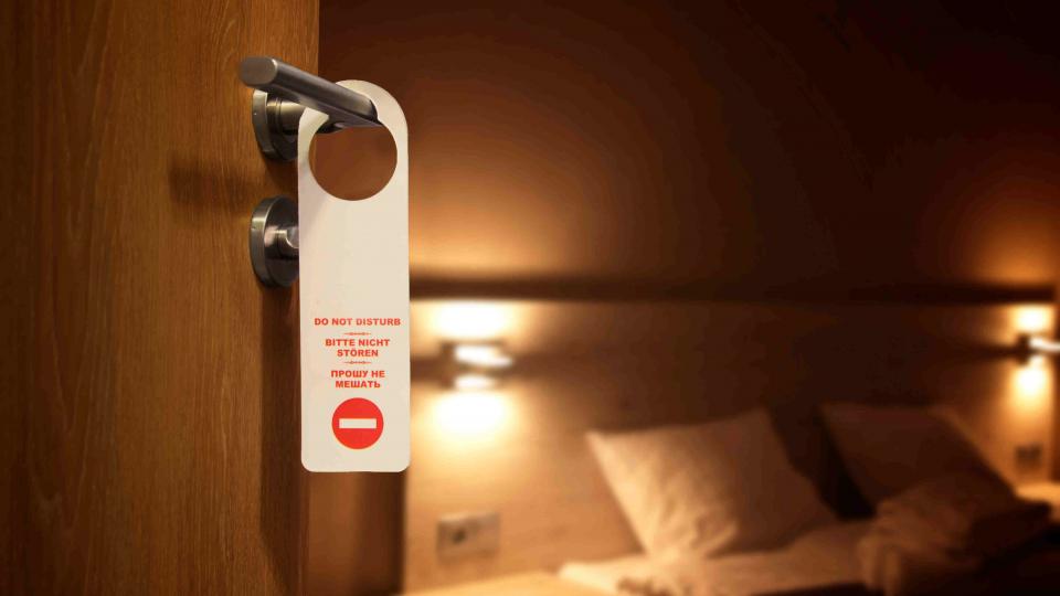 A do not disturb card on an open hotel room. There is a hotel bed in the background. 