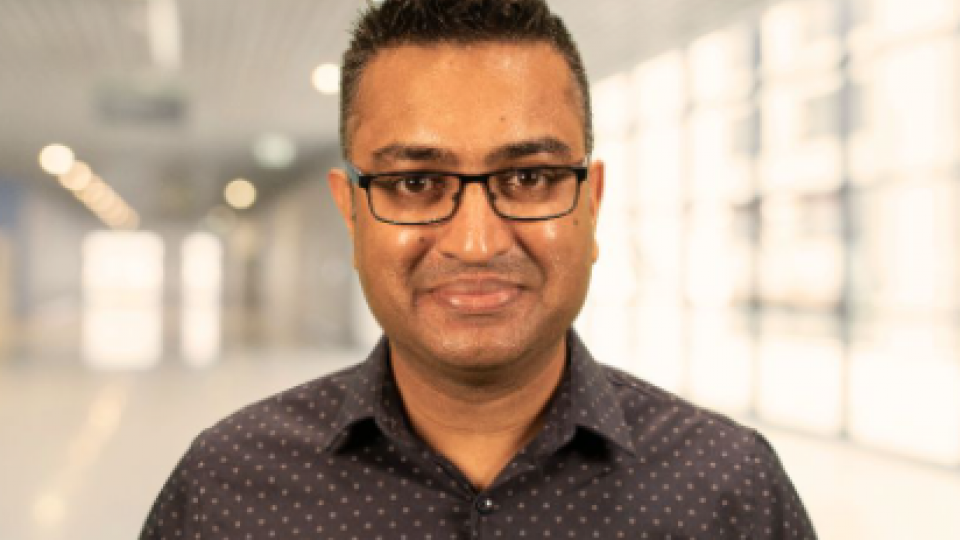 Dr Padhra has short black hair, gelled upwards and is wearing glasses whilst smiling at the camera