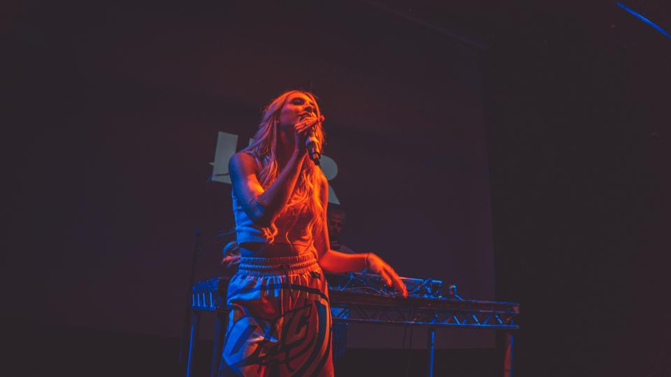 A female student performing with a microphone. She has blonde hair and is wearing a crop top and jogging bottoms. The light is red and casts dark shadows