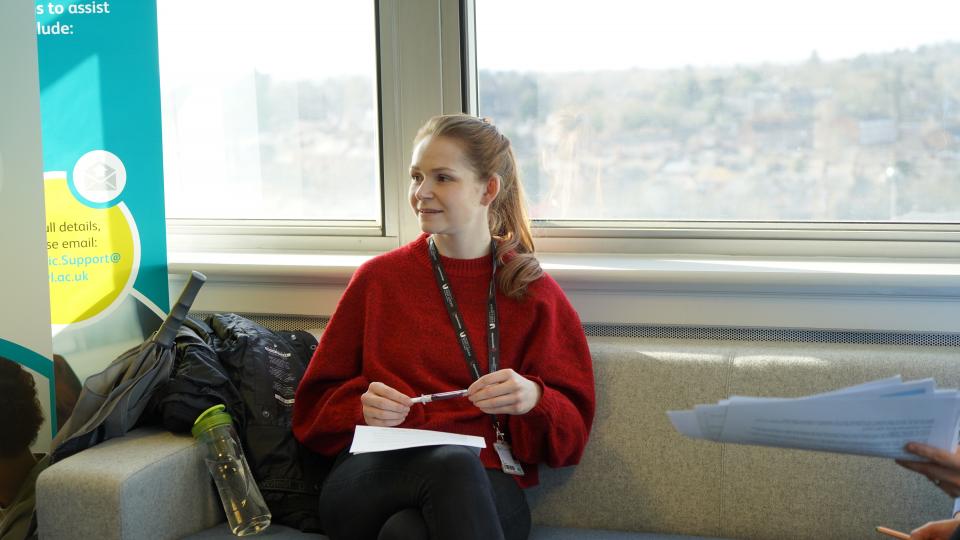 A female student with blonde hair tied back in a ponytail and a red jumper studies on a sofa in our Reading campus