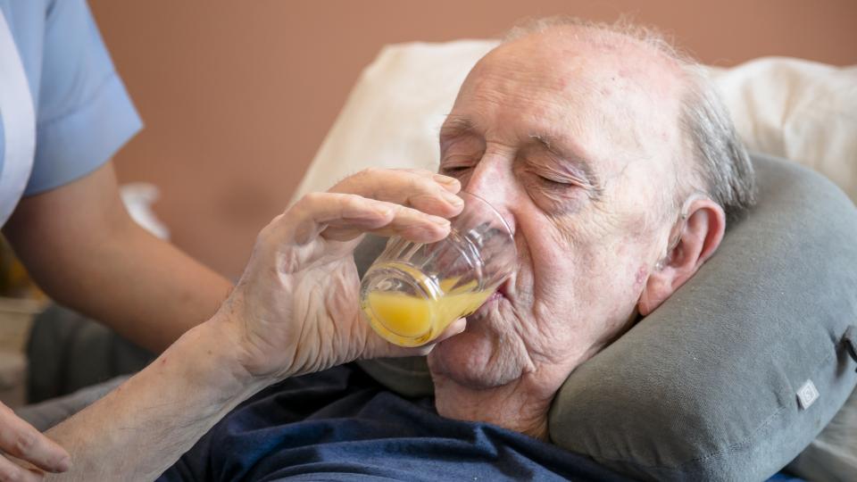 An elderly man drinking orange juice from a glass in bed.