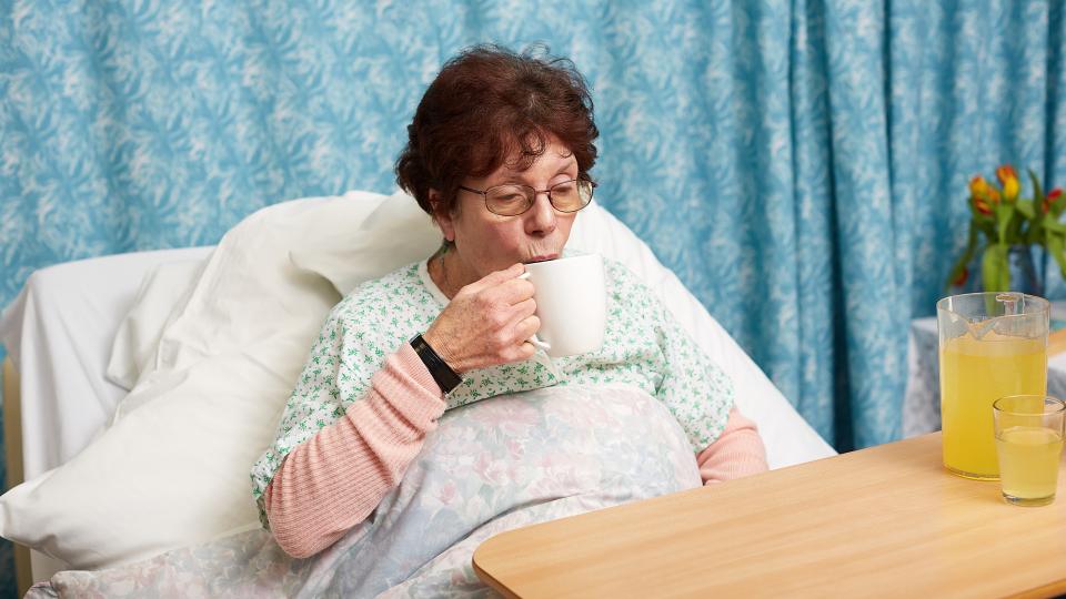 Lady in a hospital bed or care home, drinking a hot drink.