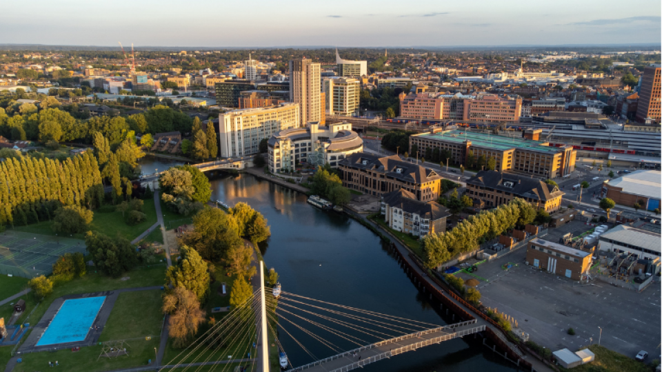 The River Thames runs through the centre of Reading Town, there is a green area on one side and some highrise buildings on the other side. The image is taken from a high perspective and includes bridges over the river. 