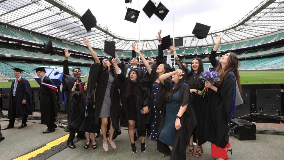 Students in gowns throwing their mortarboard hats into the air at Twickenham Stadium.
