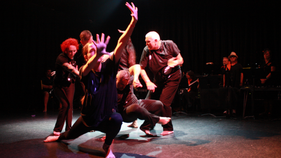 A group of people are on stage in a dynamic pose. The person closest to the camera is in a deep lunge with her arms outstretched. They are all wearing black outfits.