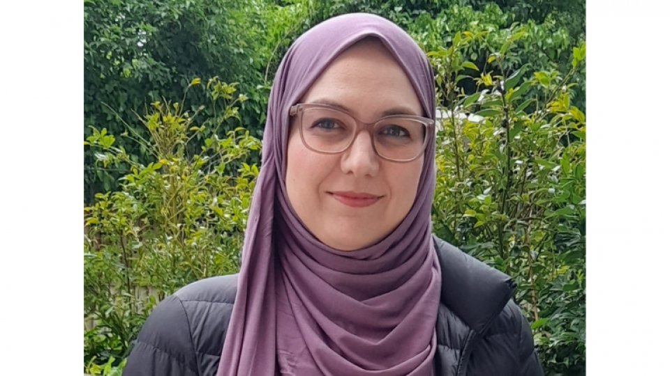Nadia is stood outside infront of some green shrubs and bushes. She is wearing a purple hijab and has rectangle glasses on and has green eyes.