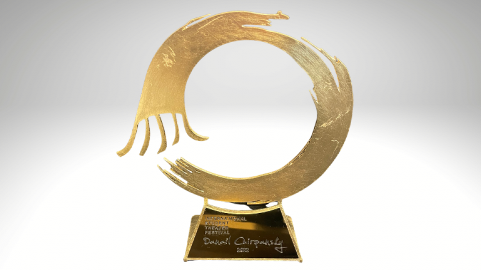 Gold trophy in the shape of an open ended circle. The circle is resting on a gold stand. The background is a grey gradient
