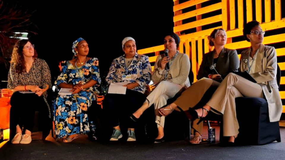 A panel of women are sat on stage all looking to the right. The stage is black with wooden pallets lit up behind them.