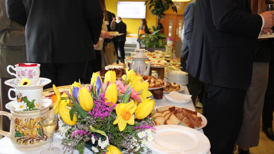 Tea cups, plates, cake and flowers presented on a table at the Donors and Scholars event with people surrounding.