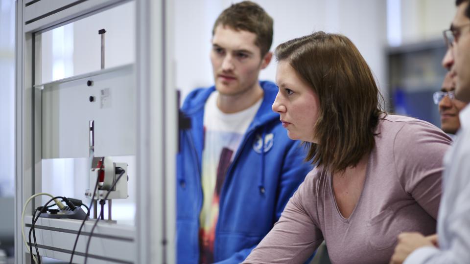 A group of students standing and working on a task together, while one is plugging something into a socket.