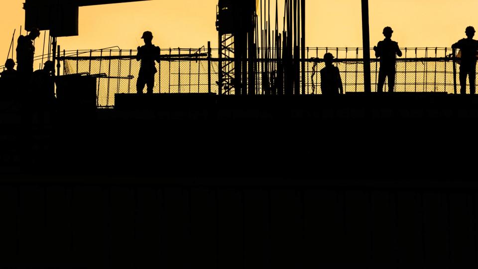 Four construction workers working on site, with a crane behind them, surrounded by yellow sky.