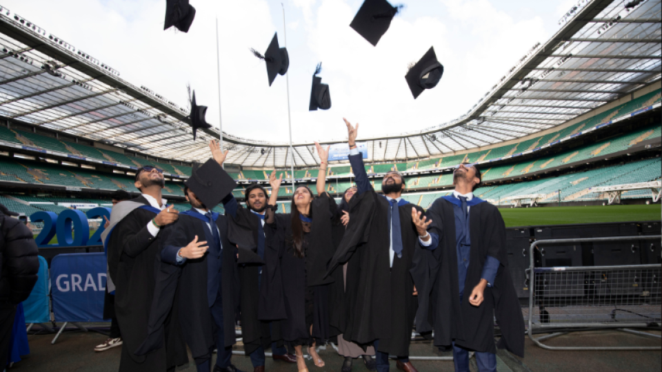 A group of graduates pitchside at Twickenham Stadium throwing their hats in the air.