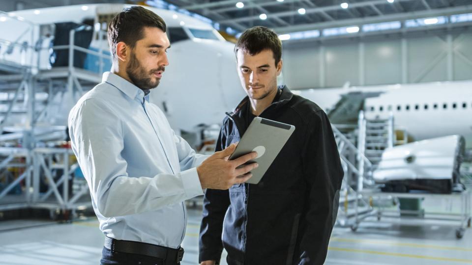 A smartly dressed man showing another man something on a tablet in front of a plane