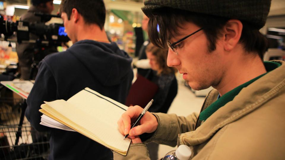 A man wearing glasses making notes behind a film crew