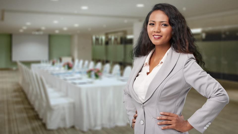 A smartly dressed woman in a conference hall in front of a long table