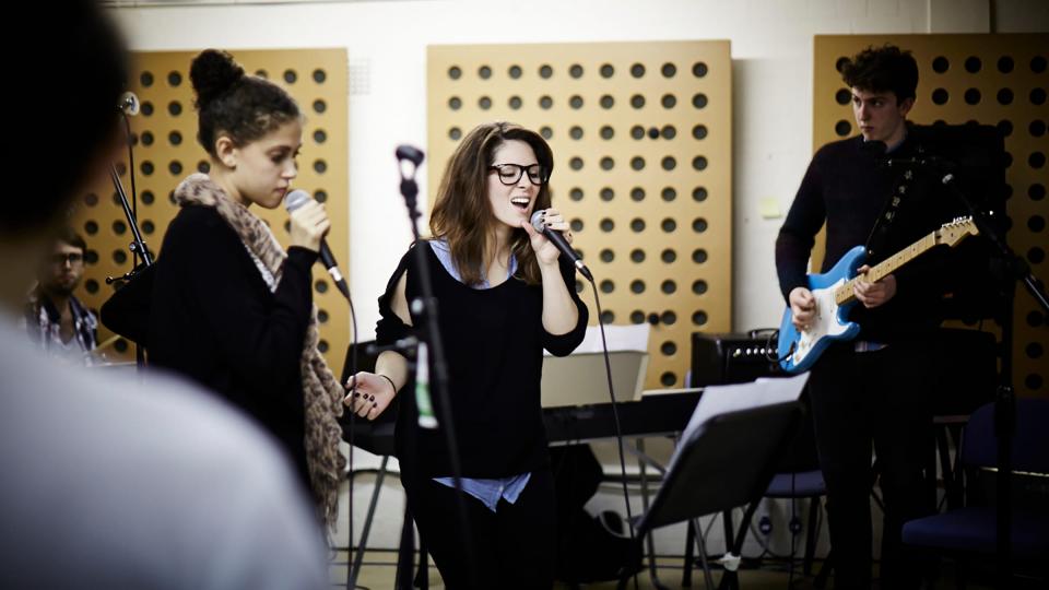 A female singing whilst a band perform in the background of a rehearsal room