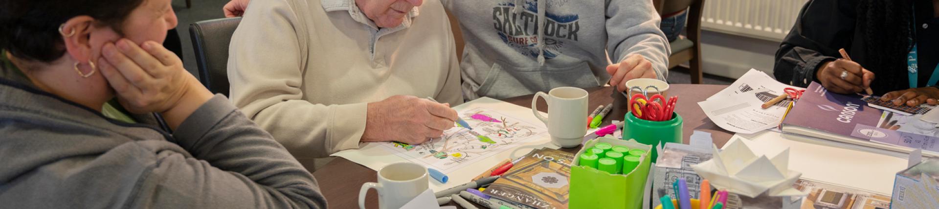 An older man is drawing using coloured pens at a table that is covered in art supplies. In the backgrounds there are other tables with different people sat at them.