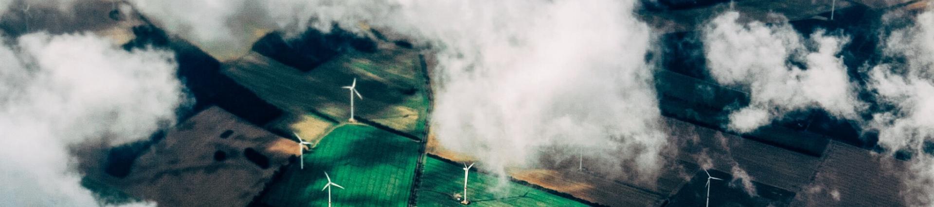 Ariel shot of green and yellow fields. Through the white fluffy clouds, you can see wind turbines dotted over the fields.