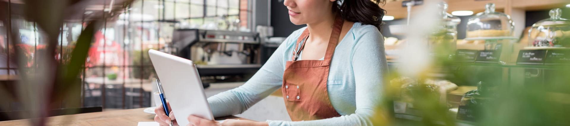A female woman wearing an apron looking at a tablet and notes in a coffee shop
