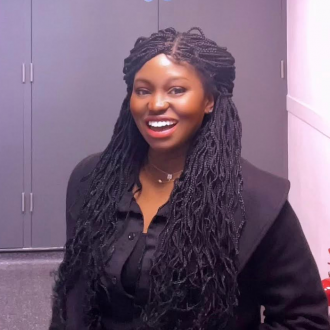 Francisca Umeh is wearing a black shirt and jacket, she has long black braids and is smiling at the camera.
