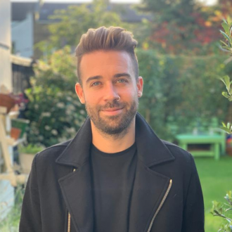 Marcus is wearing a black coat with a large collar. He is standing in a garden and has blue eyes and short brown hair. He has a short stubble beard.